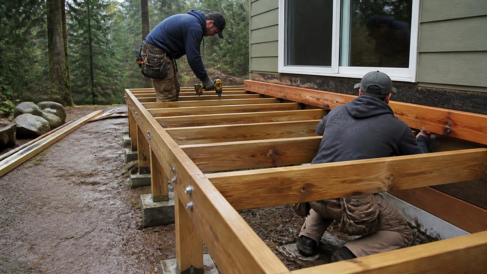 Deck building crew installing joists on a new deck frame behind a house surrounded by evergreen trees in early spring